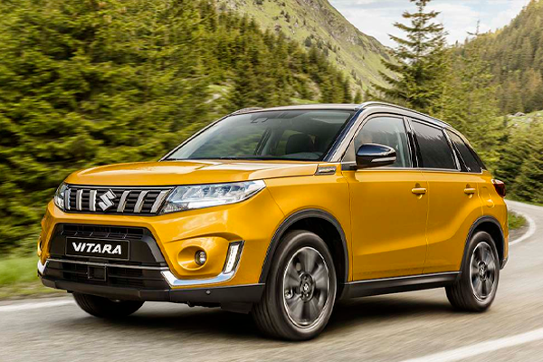 Suzuki Vitara naranja circulando por una carretera de montaña con bosque de pinos y laderas verdes al fondo