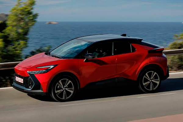Toyota C-HR rojo con techo negro circulando por una carretera de costa, con el mar y los árboles de fondo