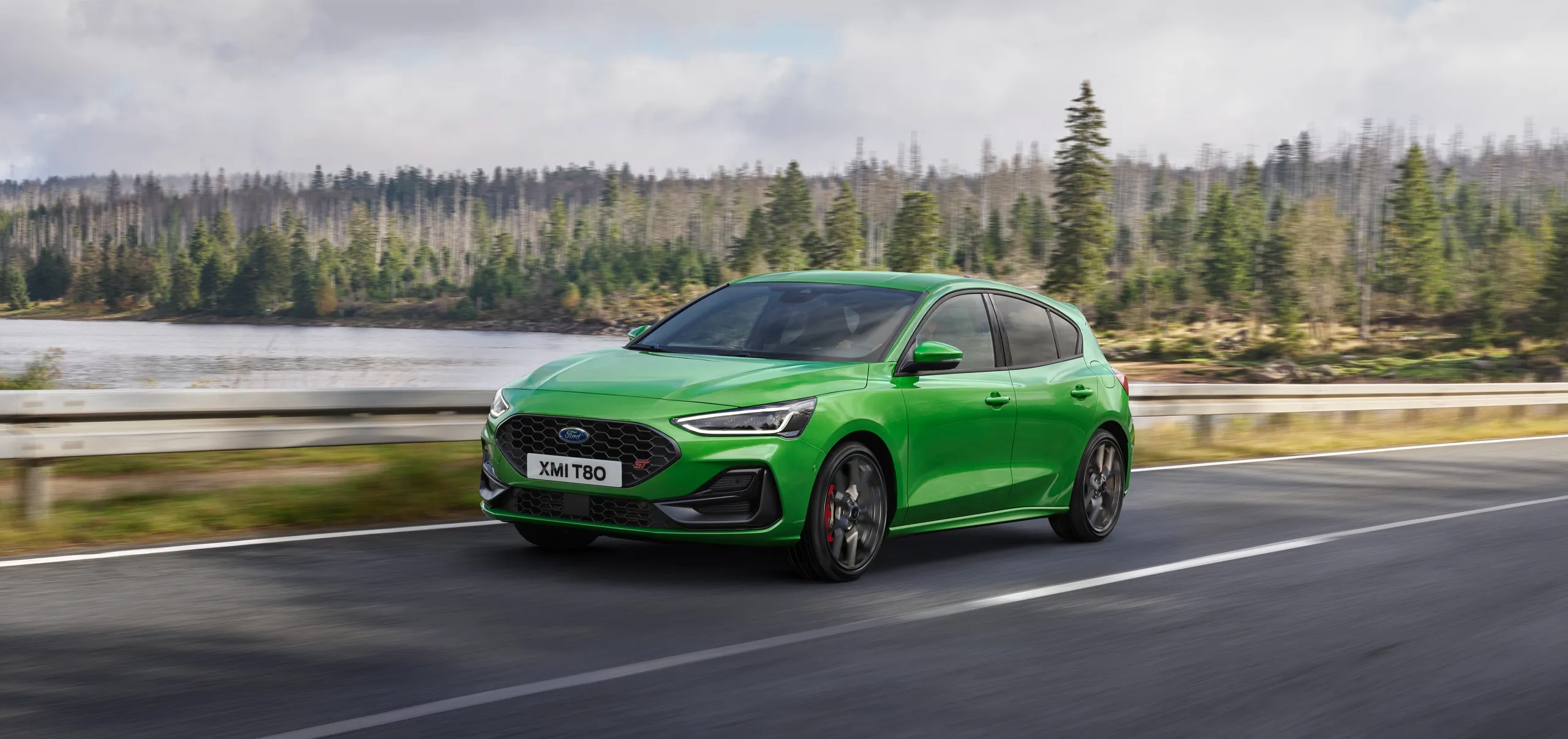 Ford Focus ST verde circulando por carretera con lago y bosque al fondo