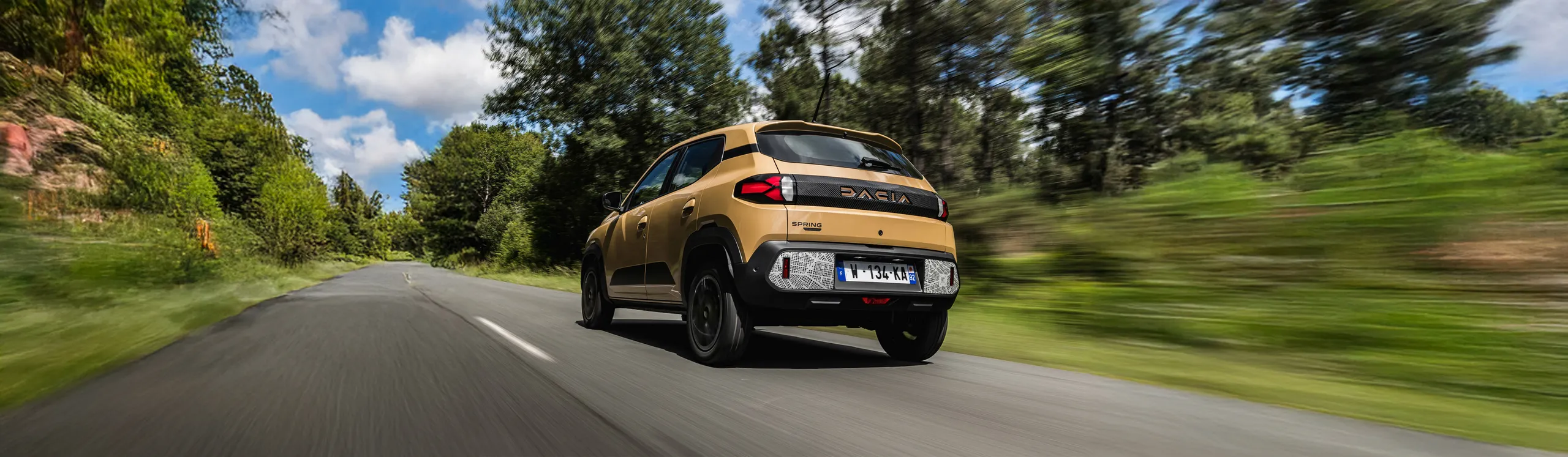 Dacia Sandero eléctrico beige circulando por carretera rural con bosque verde