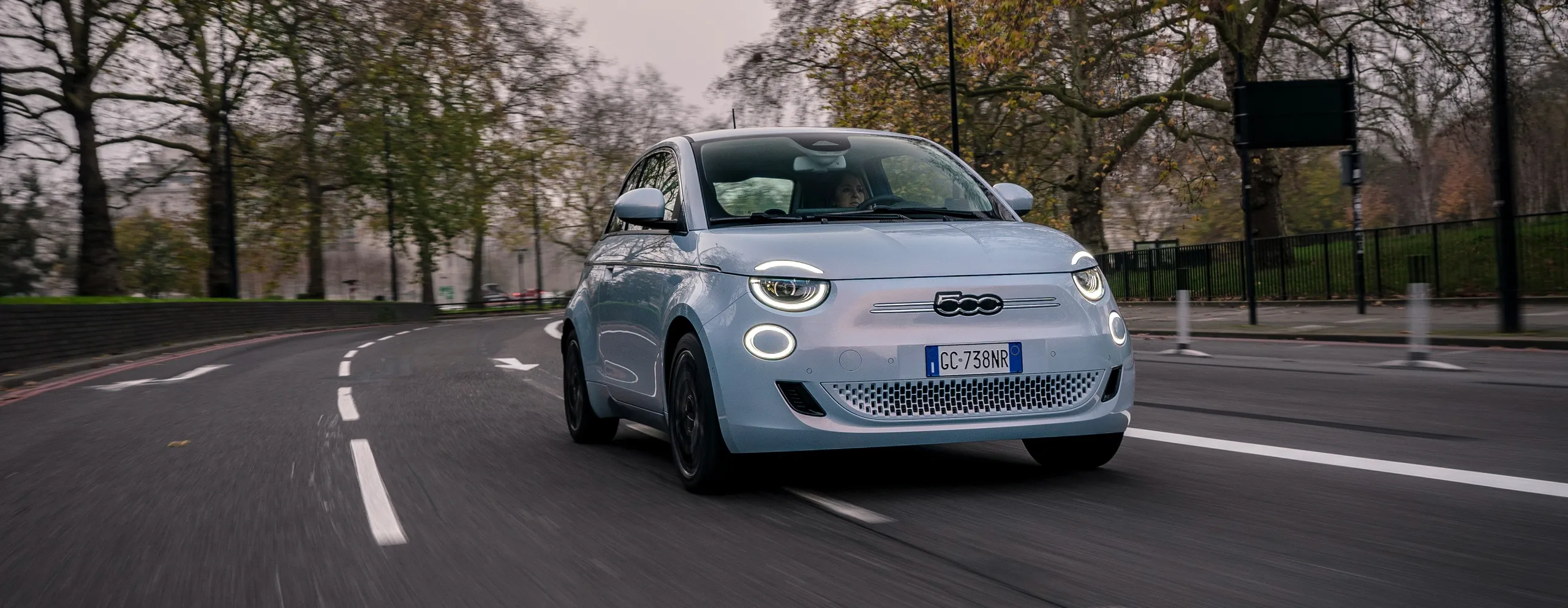 Fiat 500 eléctrico azul claro circulando por una calle urbana con árboles
