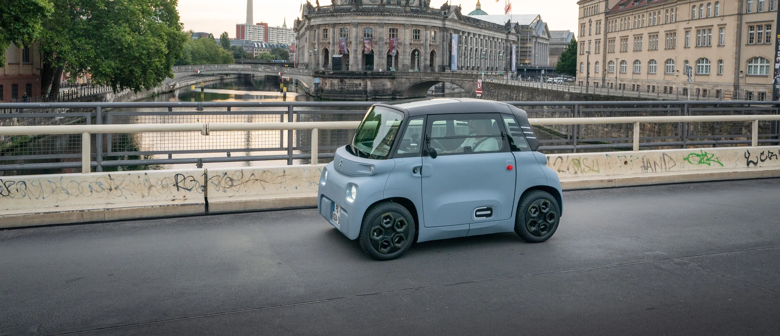 Citroën Ami azul claro circulando por un puente urbano junto a un río
