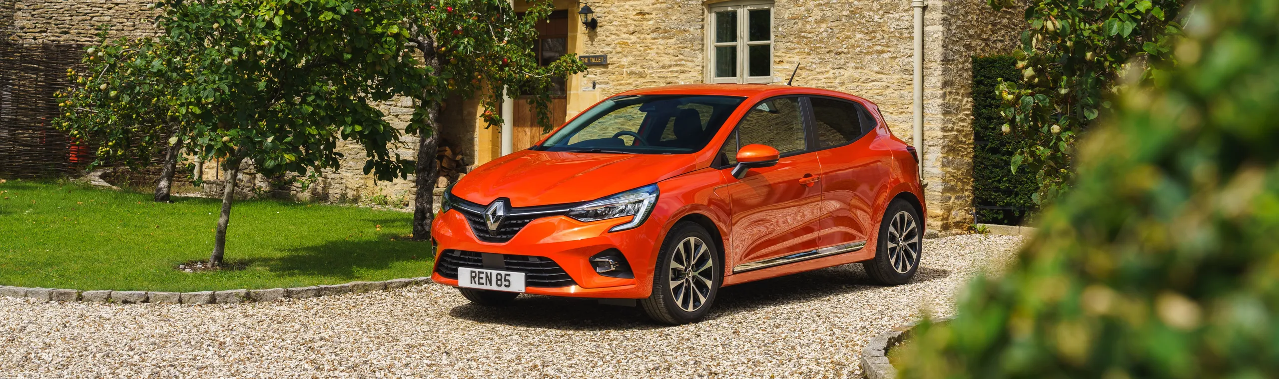 Renault Clio naranja aparcado en entrada de casa de piedra con jardín