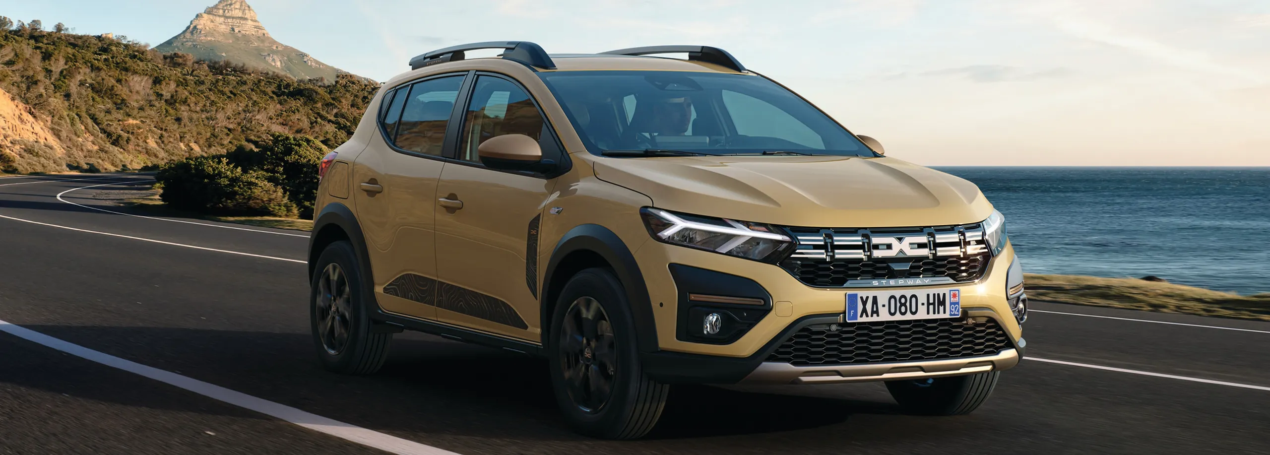 Dacia Sandero amarillo circulando por carretera costera con montaña al fondo