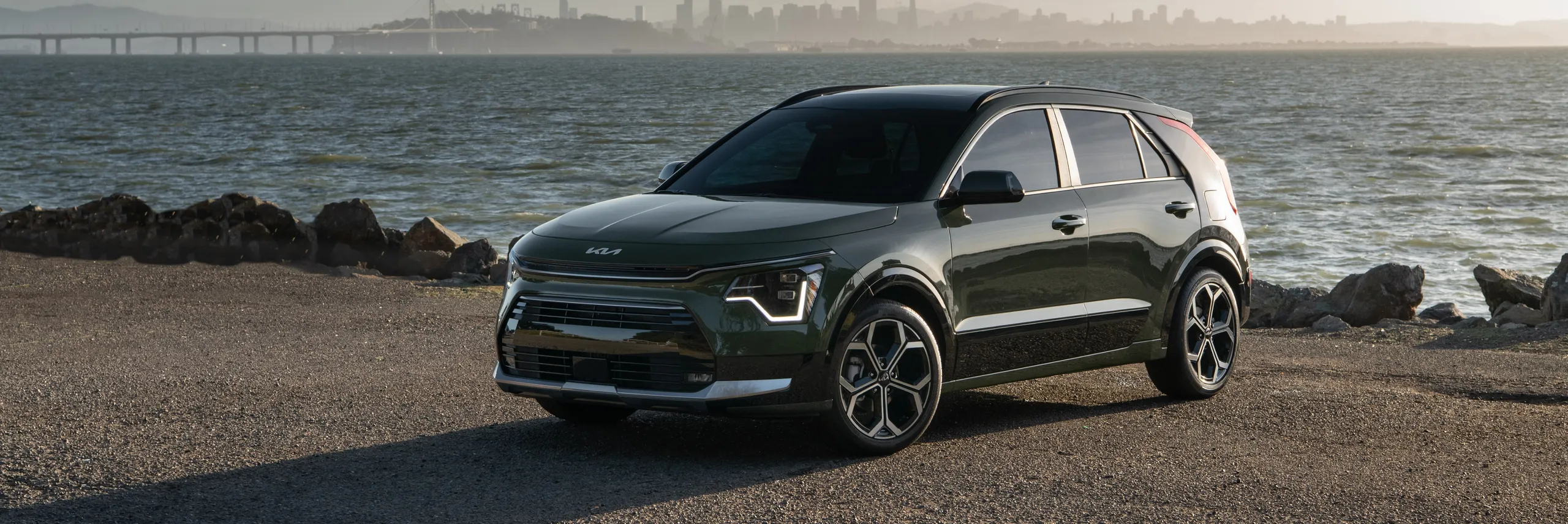 Kia Niro HEV gris oscuro estacionado en playa con ciudad al fondo
