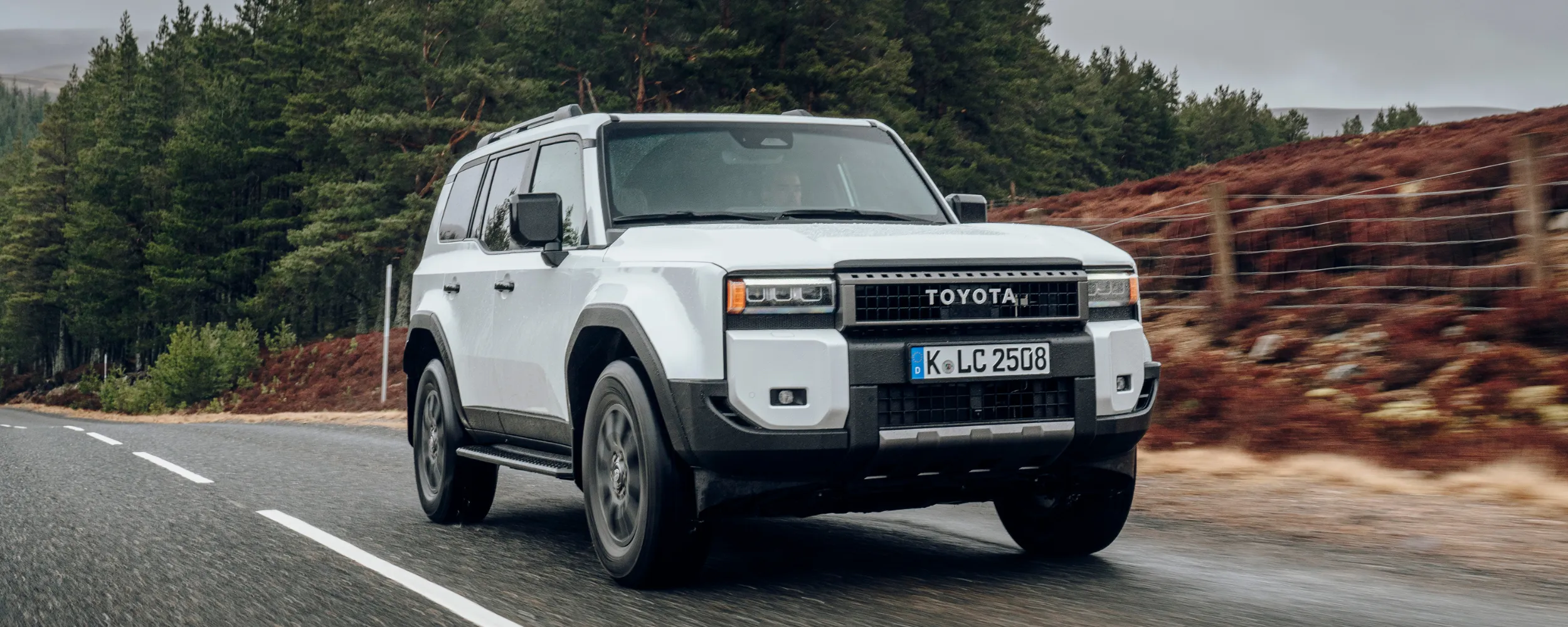 Toyota Land Cruiser blanco circulando por carretera de montaña con bosque de fondo