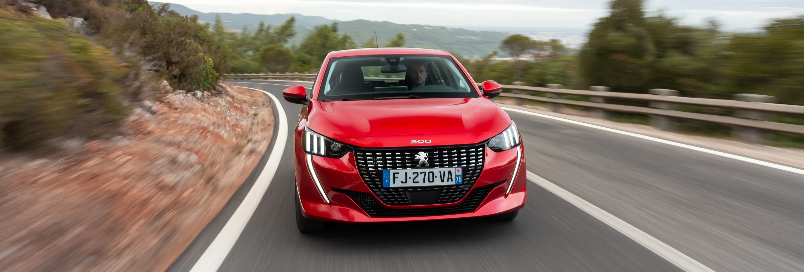 Peugeot 208 rojo circulando por autopista en curva con montañas al fondo
