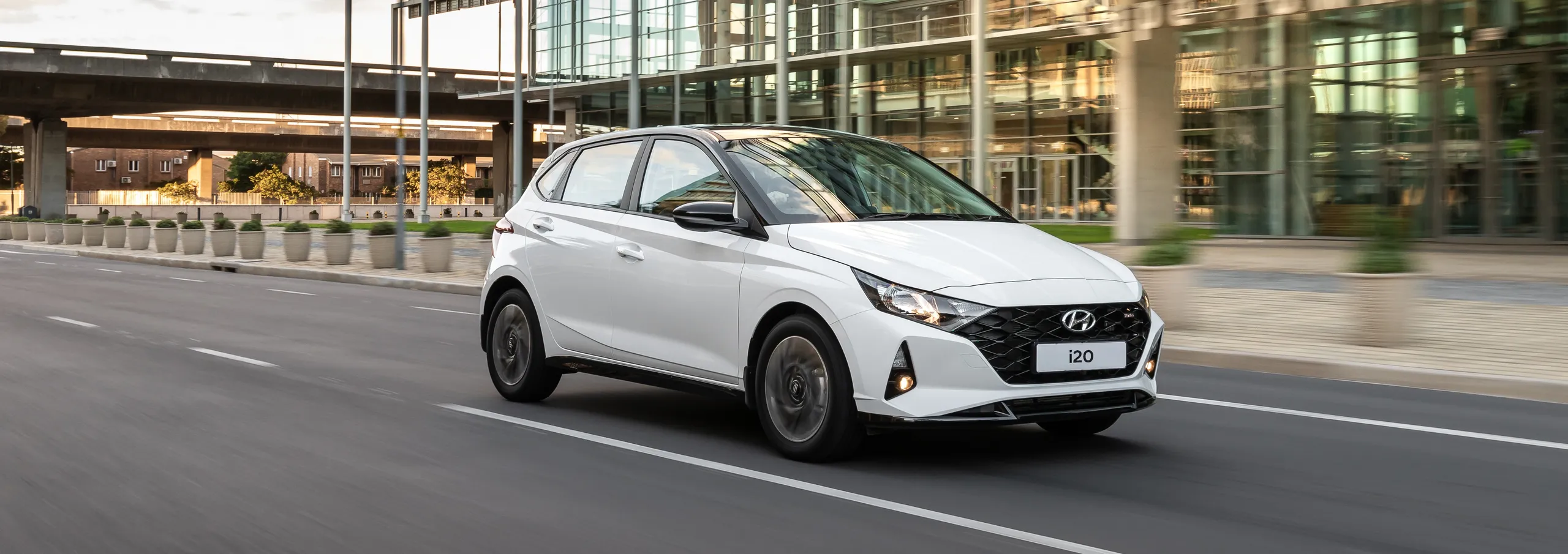 Hyundai i20 blanco circulando por calle urbana moderna con edificios de cristal