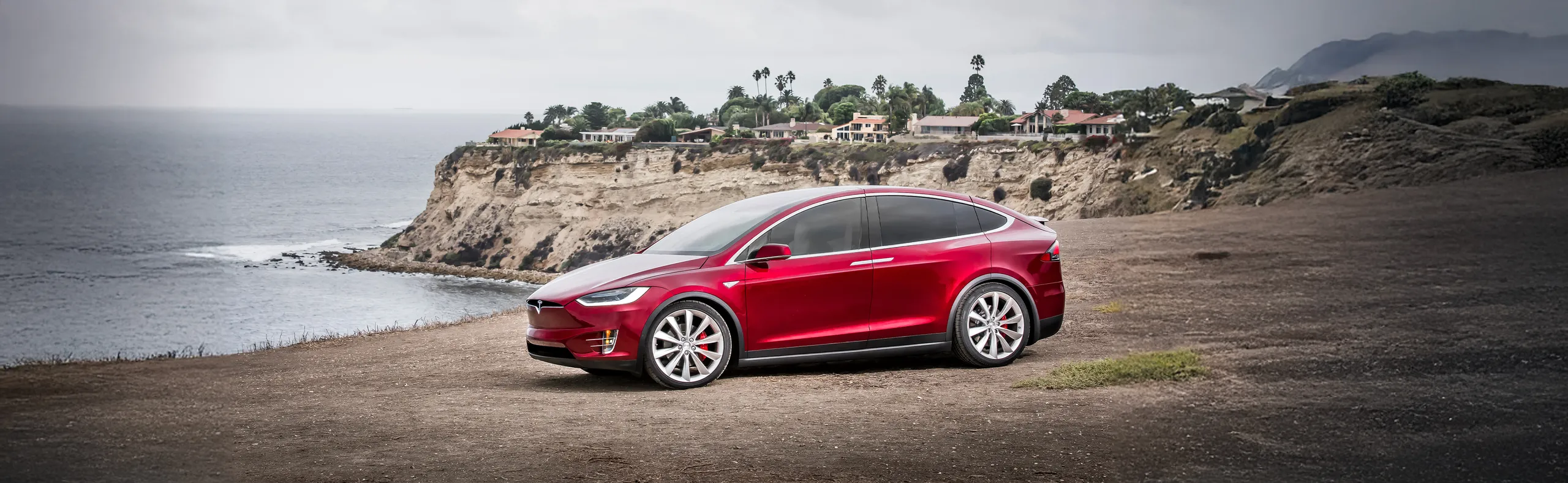 Tesla Model X rojo aparcado en acantilado frente al mar con viviendas al fondo